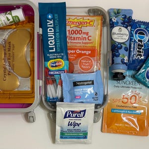May include: A clear plastic travel case containing various health and beauty items. Contents include eye masks, lip balm, hydration mix, vitamin C sachets, hand cream, chewing gum, sunscreen, and makeup remover wipes. The case is pink and has wheels.