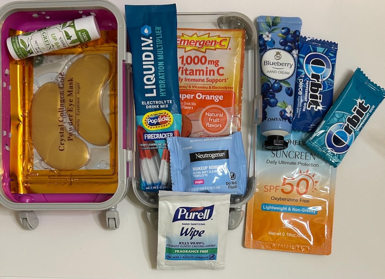 K&ouml;nnte beinhalten: Eine transparente Reisebox mit Gesundheits- und Sch&ouml;nheitsprodukten. Zu den Artikeln geh&ouml;ren Augenmasken, Lippenbalsam, Hydratisierungsmix, Vitamin-C-P&auml;ckchen, Handcreme, Kaugummi, Sonnencreme und Make-up-Entferner-T&uuml;cher. Die Box ist rosa und hat R&auml;der.
