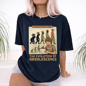 Puede incluir: Camiseta azul marino con un gráfico que dice "NATURAL HISTORY SERIES" y "THE EVOLUTION OF OBSOLESCENCE". El gráfico muestra la evolución del hombre y un niño dibujando. La camiseta tiene mangas cortas.