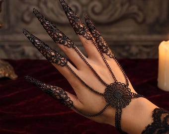 May include: A black metal hand harness featuring intricate filigree patterns on five pointed finger caps. The finger caps are connected by delicate chains to a central circular filigree medallion, which is linked to a matching wrist bracelet.