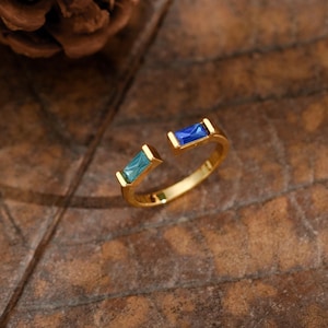 May include: A gold-toned open ring featuring two rectangular baguette-cut stones, one in a vibrant blue and the other in a lighter teal shade, set at each end of the band, creating a distinct contrast in color and a sleek, polished appearance.
