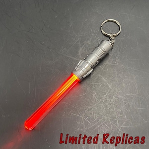 May include: A red and orange lightsaber keychain with a silver handle and a keyring. The lightsaber blade is illuminated, and the text "Limited Replicas" is visible in the image.