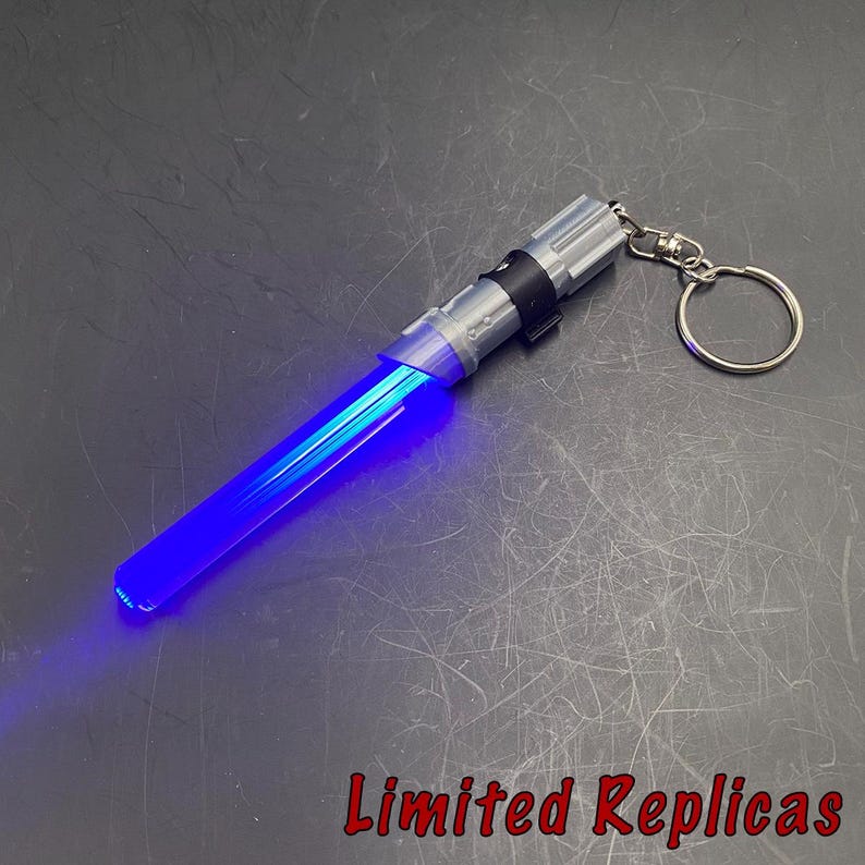 May include: A blue lightsaber keychain with a silver handle and a keyring. The lightsaber blade glows brightly. The text "Limited Replicas" is in the bottom right corner.