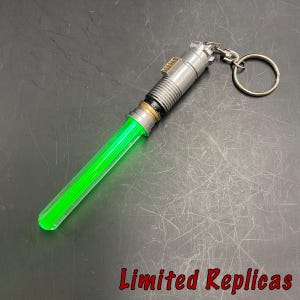 May include: A green lightsaber keychain with a silver handle and a keyring. The lightsaber blade is a vibrant green color. The text "Limited Replicas" is in red at the bottom.