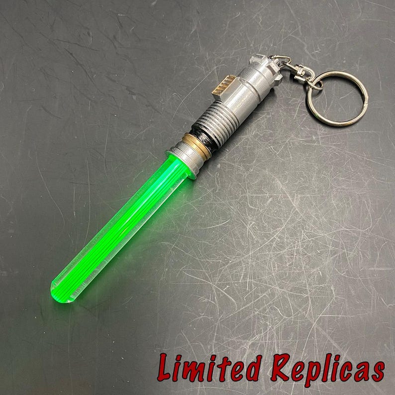 May include: A green lightsaber keychain with a silver handle and a keyring. The lightsaber blade is a vibrant green color. The text "Limited Replicas" is in red at the bottom.