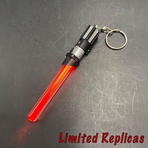 May include: A red and black lightsaber keychain with a silver key ring. The lightsaber has a red blade and a black and silver hilt. The text "Limited Replicas" is at the bottom.