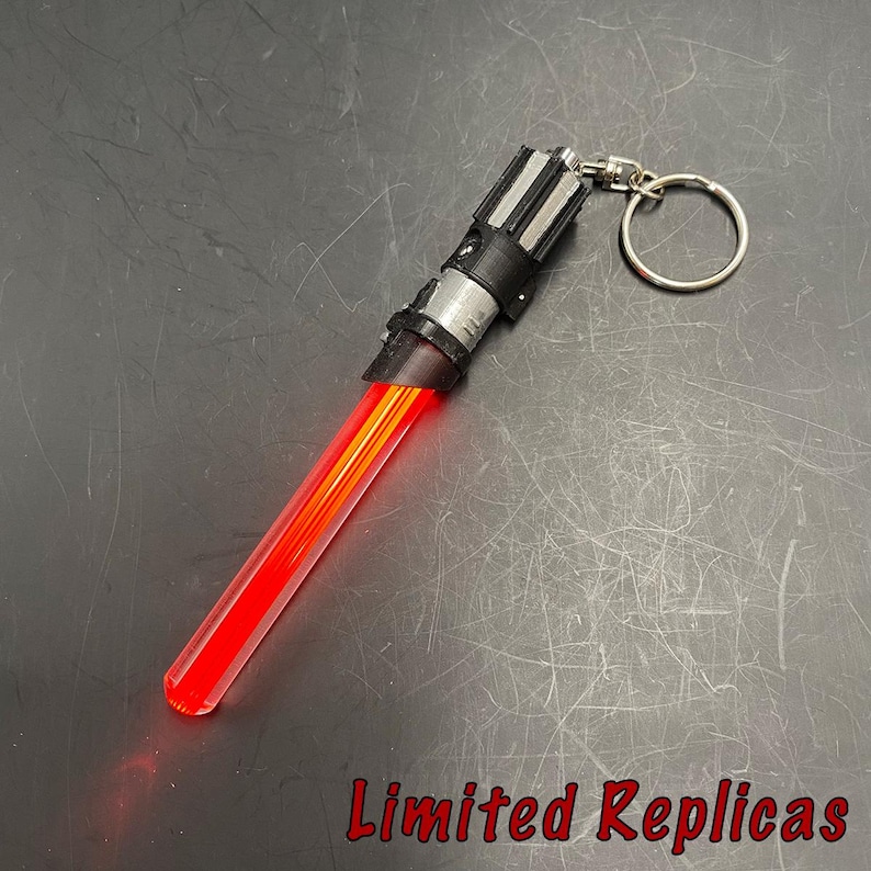 May include: A red and black lightsaber keychain with a silver key ring. The lightsaber has a red blade and a black and silver hilt. The text "Limited Replicas" is at the bottom.