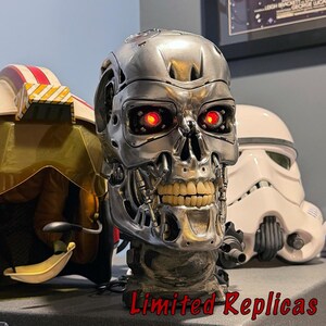 May include: A metallic silver robotic skull replica featuring glowing red eyes and visible mechanical jaw details. The skull is positioned between a pilot helmet and a white stormtrooper helmet, with the text Limited Replicas at the bottom.