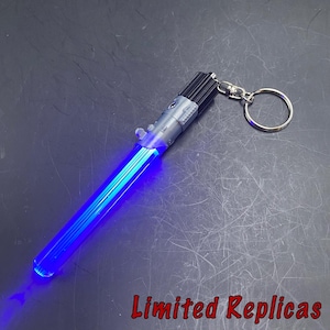 May include: A blue lightsaber keychain with a silver handle and a black emitter. The lightsaber is illuminated with a bright blue light. The keychain includes a silver keyring. The text "Limited Replicas" is visible.