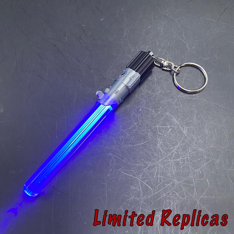May include: A blue lightsaber keychain with a silver handle and a black emitter. The lightsaber is illuminated with a bright blue light. The keychain includes a silver keyring. The text "Limited Replicas" is visible.
