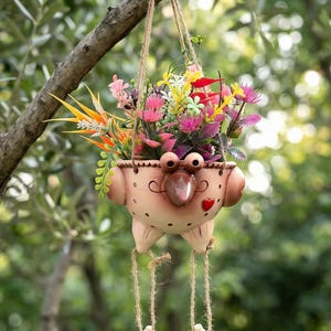 May include: A novelty hanging planter, shaped like a face with a large nose, eyes, and a red heart. The planter is filled with colourful artificial flowers and suspended by a rope. The legs of the planter dangle with decorative feet.