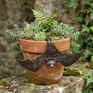 May include: A bronze-colored bat decoratively hangs on the rim of a terracotta flower pot filled with succulents and greenery. The bat has outstretched wings and detailed features. The pot sits on a stone surface, creating a natural, gothic-inspired garden accent.