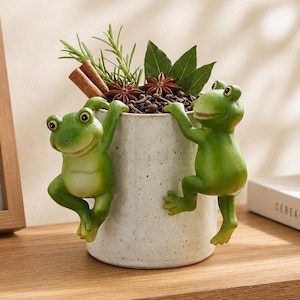 May include: A cylindrical off-white speckled ceramic planter featuring two sculpted green frogs climbing up the sides. The planter is filled with green herbs, cinnamon sticks, and star anise, creating a decorative botanical display.