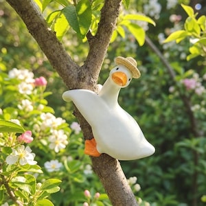 May include: A white duck figurine with an orange beak and feet, wearing a small tan straw hat. The duck is positioned as if clinging to a tree branch, with its wings spread slightly and its head tilted to the side, showcasing a smooth finish.