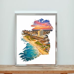 May include: A framed watercolour print of Ireland, depicting a coastal city scene within the country's outline. The artwork features a vibrant sunset, a sandy beach, and a cityscape with buildings and a tall hotel. The frame is silver, and the print is displayed on a wooden surface.