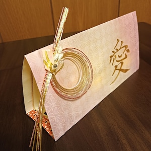 Könnte beinhalten: Ein hellrosa rechteckiger Papierumschlag mit einem dezenten geometrischen Muster und dem goldenen Kanji-Zeichen für Liebe. Er ist mit einem kreisförmigen Ornament aus goldenen und kupferfarbenen Metallkordeln, einem Papierakzent und einem Kristall verziert.