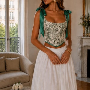 Peut inclure: Un haut corset arborant un motif toile de jouy crème et vert avec des bretelles à nouer en ruban vert foncé et un ourlet pointu, associé à une longue jupe blanche fluide dotée d'une taille froncée et d'un tissu léger et texturé.