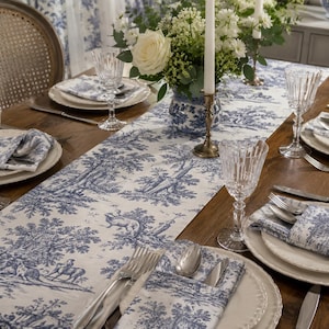 Peut inclure: Un chemin de table et des serviettes assorties arborant un motif toile de Jouy bleu et blanc avec des scènes pastorales. L'ensemble est disposé sur une table en bois avec des assiettes blanches, des verres texturés et des couverts en argent.
