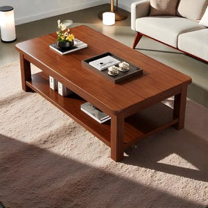 Peut inclure: Une table basse rectangulaire en bois brun foncé avec une étagère inférieure. La table a des bords arrondis et est ornée de livres, d'un plateau avec des tasses à thé et d'une petite composition florale. La table repose sur un tapis brun clair.