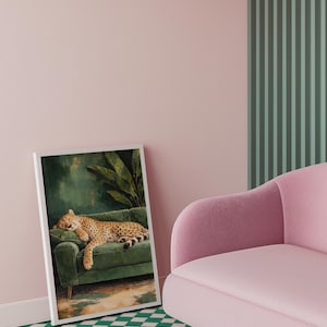 May include: A framed art print depicts a cheetah resting on a green velvet sofa. The cheetah has a spotted coat and is lying down. The background of the print is a dark green with a large leaf. The print is leaning against a pink wall.