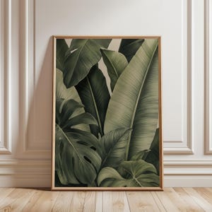 May include: A rectangular wall art print in a light wood frame featuring a detailed illustration of overlapping tropical leaves in various shades of dark and muted green. The artwork displays large banana leaves and monstera foliage.