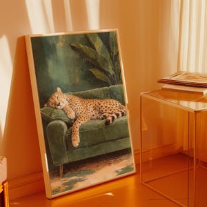 May include: A framed art print depicts a cheetah resting on a green velvet sofa. The cheetah has a spotted coat and is curled up on a cushion. The background features a green wall and tropical foliage.
