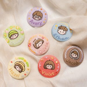 May include: Six circular fabric badges featuring embroidered cartoon faces and names. The badges are lavender with Avery, light green with Oliver, pink with Olivia, light blue with Ethan, yellow with Hazel, red with Violet, and brown with Jacob.