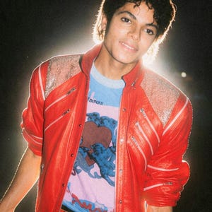 May include: A bright red leather jacket with silver zippers and textured shoulder panels, worn over a light blue t-shirt featuring a graphic of a brown heart and blue figures, paired with black trousers and a brown leather belt with a gold chain.
