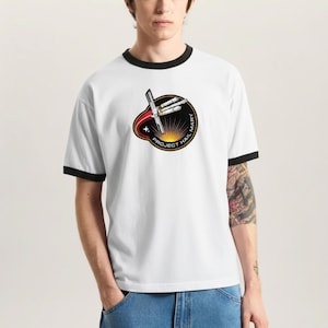 May include: White ringer t-shirt with black trim featuring a circular graphic. The graphic includes a spaceship design, the text "PROJECT HAIL MARY", and a sunburst design in red, yellow, and black.
