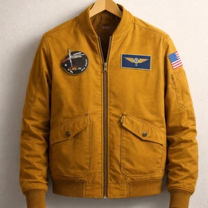 May include: Mustard yellow bomber jacket with a zip-up front, two front pockets, and ribbed cuffs and hem. Features patches including an American flag and a military-style emblem. The jacket is hanging on a wooden hanger.