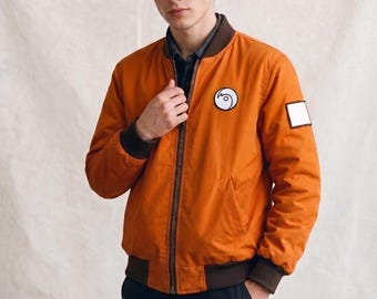 Peut inclure: Un blouson aviateur orange avec un col, des poignets et un ourlet côtelés marron foncé. La veste présente une fermeture éclair, un écusson blanc circulaire avec un motif en spirale noire sur la poitrine gauche et un écusson blanc rectangulaire.