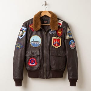 Op de afbeelding: Bruine leren bomberjack met een schapenvachtkraag en diverse geborduurde patches. De jas heeft twee voorzakken en geribde manchetten en zoom. Patches tonen militaire en luchtvaart gerelateerde ontwerpen.