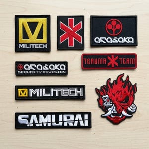 May include: A collection of embroidered patches with various designs and text. The patches feature the words "MILITECH", "ARASAKA", "TRAUMA TEAM", and "SAMURAI". The patches are black with white, yellow, and red accents.