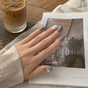 Puede incluir: Un conjunto de uñas manicuradas con un acabado brillante en tonos azul pizarra y una uña de acento en color blanco roto. La uña del pulgar muestra un detallado patrón floral blanco sobre un fondo azul pizarra, creando un aspecto uniforme.