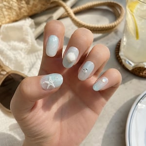 Può includere: Unghie di forma ovale con una base sfumata in azzurro e bianco. Le unghie sono decorate con applicazioni bianche tridimensionali, tra cui una piccola stella marina, una conchiglia, minuscole perline argentate e una singola perla iridescente.