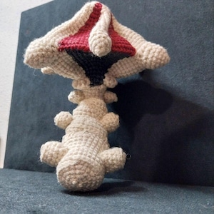 May include: A handmade crochet mushroom in cream, red, and black. The mushroom has a detailed, textured design with a rounded cap and a stem with small, rounded protrusions. The mushroom is displayed against a dark background.