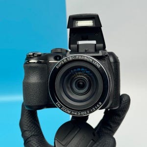 May include: A black digital camera with a pop-up flash and a Super EBC Fujinon lens. The lens barrel features text reading 24x ZOOM f=4.3-103.2 1:3.1-5.9. The camera body has a textured grip on the left side and a circular lens cap below the main body.