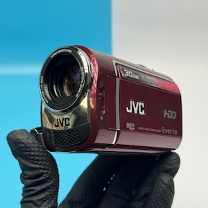 May include: A deep red JVC Everio hard disk camcorder featuring a Konica Minolta lens, 30x optical zoom, and 30GB storage capacity. The device includes a Micro SD card slot, HDD branding, and clear white text labels on the side panel.