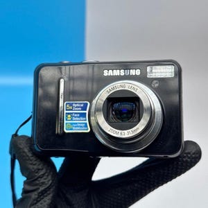 May include: A black Samsung digital camera featuring a silver lens with 6.3-31.5mm zoom capabilities. The front body displays a Samsung logo, a flash unit, and a sticker highlighting 5x optical zoom, face detection, and digital image stabilization.