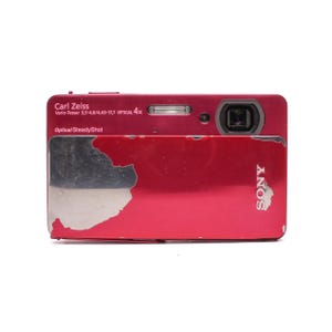 May include: A red Sony digital camera with a sliding front cover. The camera features a Carl Zeiss Vario-Tessar 3,5-4,6/4,43-17,7 Optical 4x lens and Optical SteadyShot technology. The red finish on the sliding cover shows significant wear and paint loss.