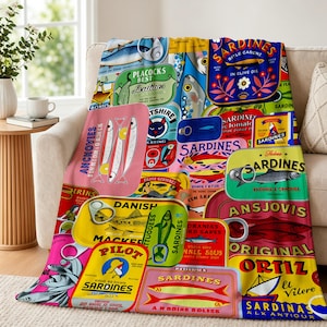 May include: A colorful throw blanket featuring a collage of vintage sardine can labels. The labels are in various shapes and colors, with text and illustrations of sardines. The blanket is draped over a beige sofa.