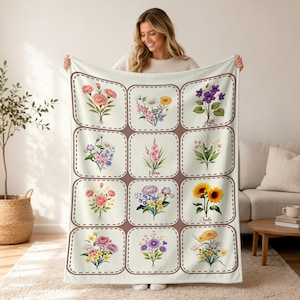 May include: A cream-colored blanket featuring a grid of floral illustrations in various colors. Each square contains a different bouquet with names like "Hall", "Nora", and "Jude". The blanket is held up by a person.