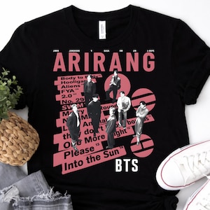 Puede incluir: Camiseta negra con un diseño gráfico rosa con la palabra "ARIRANG" y el texto "BTS". El diseño incluye imágenes de personas y otros textos. La camiseta se combina con vaqueros y zapatillas blancas.