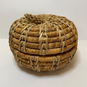 May include: A round, woven basket with a lid. The basket is made of natural-colored reeds and has a tightly woven pattern. The lid is secured with a small, twisted handle. The basket is likely used for storage or decoration.