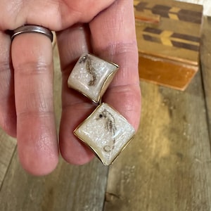 May include: A pair of square earrings with a gold-colored border. Each earring features a seahorse encased in a clear, glittery resin. The earrings are held in a hand, with a wooden box in the background.