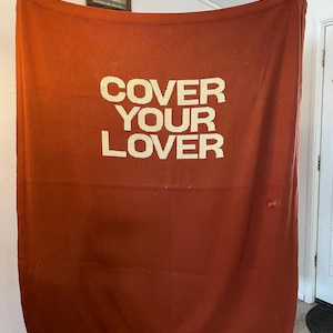 May include: A rust-colored blanket with the text "COVER YOUR LOVER" in large, off-white letters. The blanket is held up, displaying its full rectangular shape. The fabric appears to be a soft, woven material.
