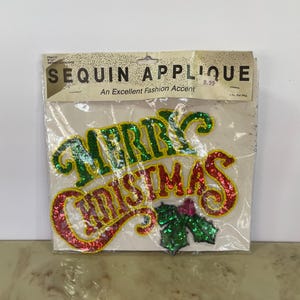 May include: A sequin applique in a clear plastic package. The applique spells "MERRY CHRISTMAS" in green and red sequins with a yellow border. The package reads "SEQUIN APPLIQUE" and "An Excellent Fashion Accent".