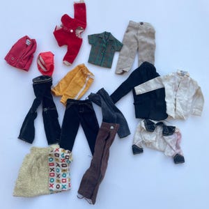 May include: A collection of vintage doll clothes in various colors and styles. Includes red, black, yellow, and brown pants, jackets, and shirts. There is also a red hat and a gold and patterned skirt.