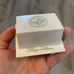 May include: A white jewelry box with a silver starburst design on the lid. The box has a rectangular shape and the words "Shifrin-Willens" are written in cursive on the base.