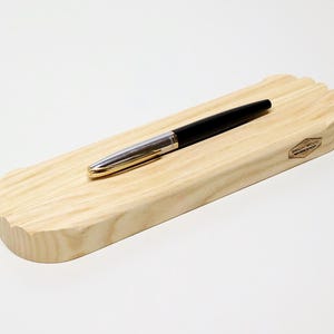 May include: A black and silver pen rests on a light-colored wooden pen holder. The pen holder is rectangular with a curved edge and a small logo. The pen has a gold-colored tip.